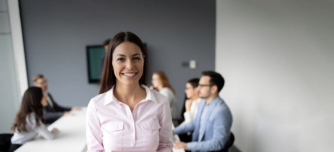 Woman wearing a pink blouse smiling with people sitting around a conference table in the background